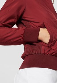 Bordeaux bomberjakke med en glat finish, ribstrikkede manchetter og en tekstureret sidelomme. Har lynlås foran og sidelomme.