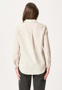 Beige langærmet skjorte med struktureret krave, knaplukning ved ærmerne og rygplissering. Glat stof med en subtil glans.