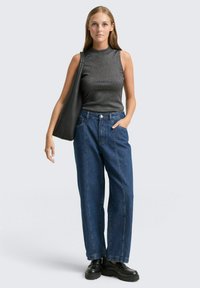 Schwarzes ärmelloses Oberteil, blaue hochtaillierte, weit geschnittene Jeans, hält eine schwarze Tragetasche. Glatte Textur, einfaches Design, lässige Passform, minimale Details.