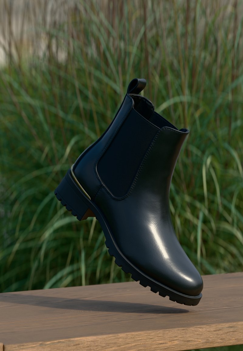 Bottine en cuir noir avec panneaux latéraux élastiques et semelle robuste, posée sur une surface en bois avec un fond d'herbe verte.
