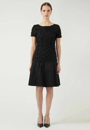 Donna in piedi che indossa una maglia nera a maniche corte con texture e una gonna nera al ginocchio, con tacchi alti neri, su uno sfondo semplice.