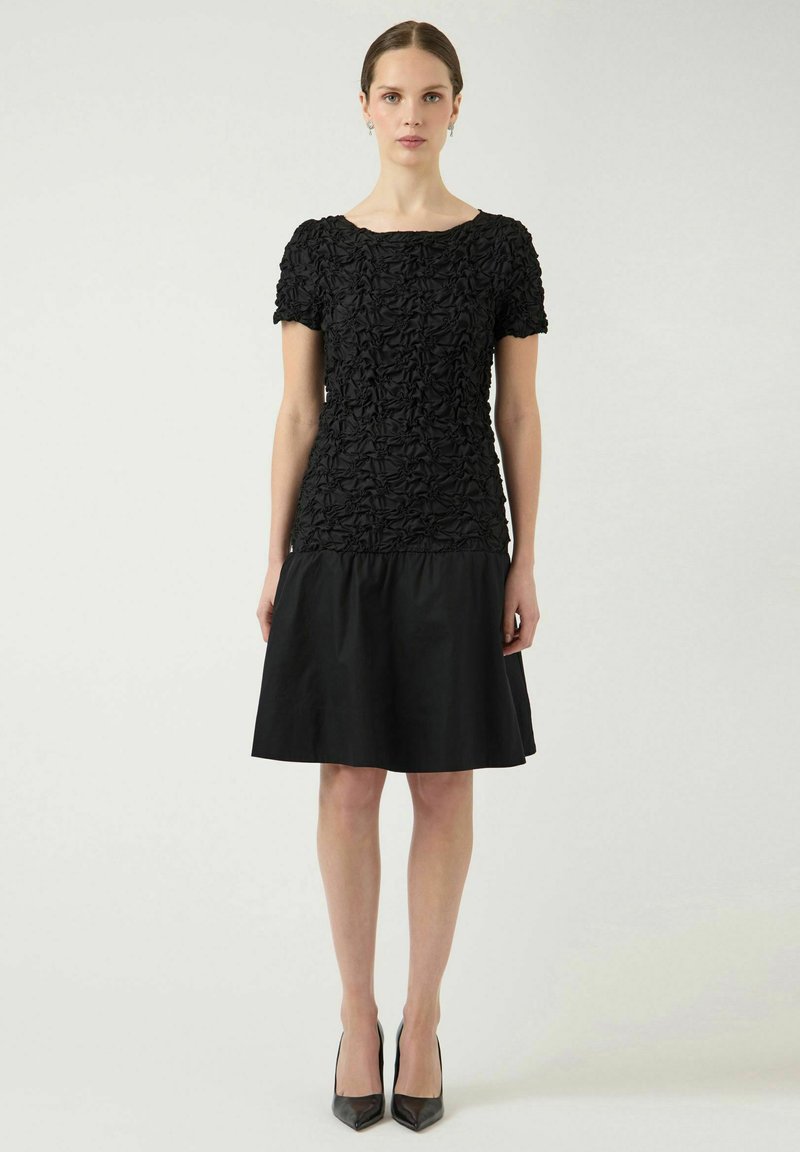 Naine seisab musta tekstuuriga lühikeste varrukatega topi ja põlveni ulatuva musta seelikuga, kandes musti kõrgeid kontsi, taust on neutraalne.