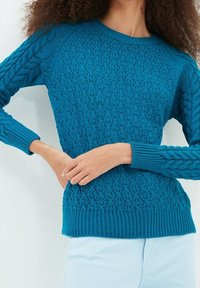 Stickad tröja i livlig teal, med texturerade kabelmönster på ärmarna, ribbade muddar och fåll, samt en figurnära passform.