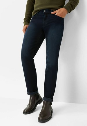 Dark blue denim jeans with a slim fit, featuring a rolled cuff, five-pocket design, and a button closure at the front. Paired with brown boots.