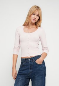 Νεαρή γυναίκα με ξανθά μαλλιά που φοράει ανοικτό ροζ μπλουζάκι με κουμπιά μακριάς μανίκας και μπλε τζιν, πόζαρε με το ένα χέρι στην τσέπη μπροστά σε λευκό φόντο.