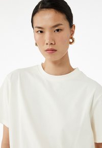 Valkoinen puuvillapaita, jossa on pyöreä kaula-aukko, lyhyet hihat ja sileä pinta. Mallilla on päällään suuret pyöreät kultaketjut.