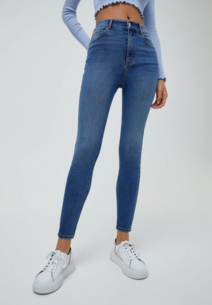 Jean bleu taille haute, coupe slim, porté avec des baskets blanches à plateforme et un haut bleu clair à manches longues, court, laissant voir le ventre et les jambes.