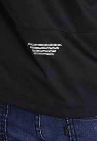 Sort atletisk jakke med en glat tekstur. Har et reflekterende accent med fem vandrette linjer på siden.