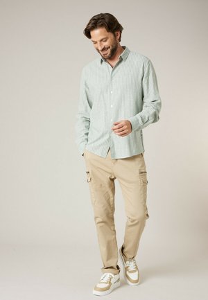 Man die lacht, draagt een lichtgroen gestreept overhemd met knopen, beige cargobroek en beige-witte sneakers, staand tegen een effen lichte achtergrond.