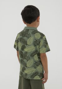 Camicia verde a maniche corte con un motivo di foglie e piante. Texture morbida, colletto polo e vestibilità comoda. Indossata con pantaloncini verdi.