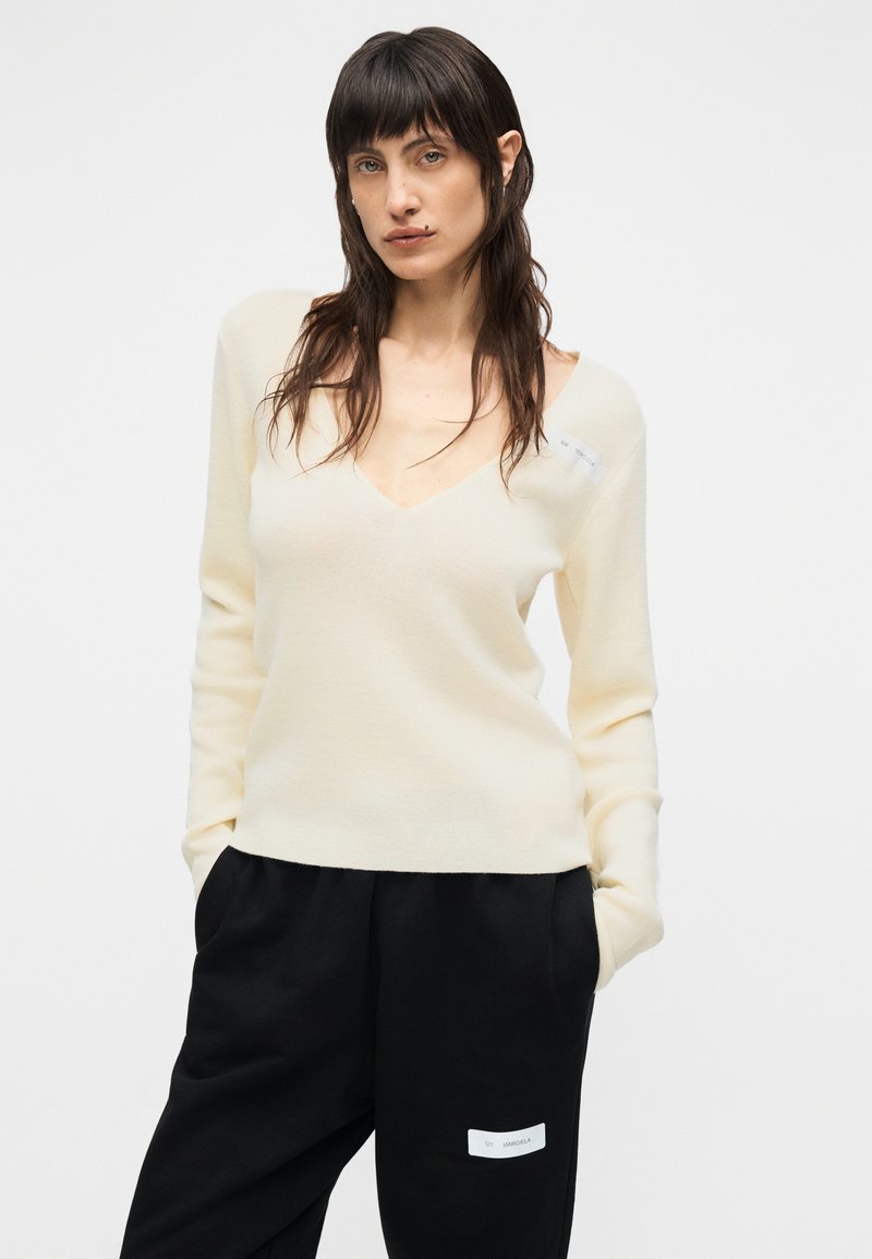 Pull en maille couleur crème avec un col en V profond, des manches longues et une coupe décontractée, associé à un pantalon noir à jambes larges avec une étiquette.