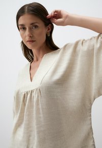 Top beige chiaro con scollatura a V e dettagli plissettati, realizzato in tessuto strutturato. Presenta maniche larghe e una vestibilità rilassata.