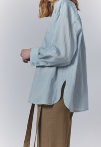 Světle modrá pruhovaná dlouhá košile s volným střihem, s zaobleným lemem a manžetami na knoflíky. V kombinaci s béžovými kalhotami s vysokým pasem.