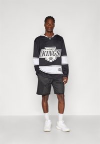 Must musta vormi "Los Angeles Kings" logoga must jääkohvri jakk, millel on valged ja hõbedased triibud, koos mustade võrkpükste ja valgete sportlike kingadega.