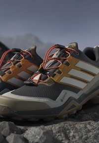 Παπούτσια trail running με πορτοκαλί κορδόνια, κίτρινες πλαϊνές ρίγες και ετικέτα Gore-Tex σε βραχώδες υπαίθριο χώρο κάτω από καθαρό ουρανό.