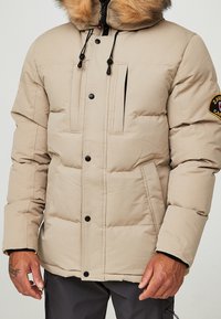 Chaqueta acolchada beige con capucha de pelo, que cuenta con un cierre frontal de cremallera y broches, dos bolsillos laterales y un pequeño parche con logo en la manga.