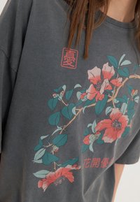 Szary bawełniany T-shirt z kwiatowym nadrukiem, na którym widnieją różowe i turkusowe kwiaty, zielone liście oraz czerwone akcenty. Luźny krój.