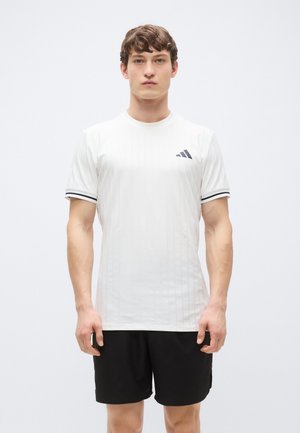 White athletic shirt with vertical stripes, black collar and sleeve accents, featuring a small black logo on the chest. Worn with black shorts.