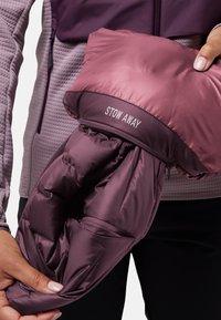 Handen die een opgevouwen paarse jas vasthouden met de tekst "STOW AWAY", gedragen over een gestructureerde lichtpaarse zip-up top en zwarte broek.