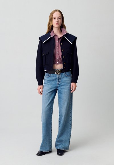 Chaqueta de lana recortada en marino con un collar oversize, combinada con una camisa de cuadros roja. Jeans de pierna ancha en azul claro y zapatos negros completan el conjunto.