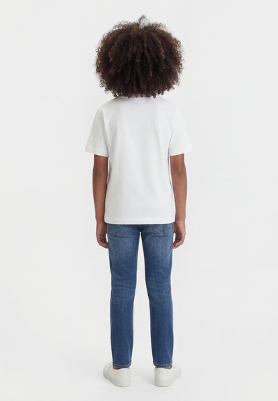 Otrok z kodrastimi lasmi, oblečen v enostavno belo majico, modre jeans hlače in bele superge, stoji obrnjeno stran proti svetlemu preprostem ozadju.