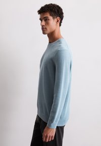 Pull en maille texturée bleu clair avec un col rond et des manches longues, associé à un pantalon sombre. Design minimaliste sans logos visibles.