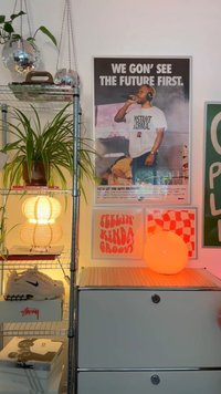 Metallhylla med växter, en trä-lampa och olika konsttryck. En sfärisk orange lampa och Nike-skor på en ljusgrå byrå.