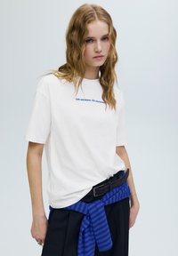 Biała bawełniana koszulka z niebieskim napisem „Off-season. On purpose.” Włosy zaczesane do tyłu, ciemny pasek i niebieski sweter w paski związany w talii.