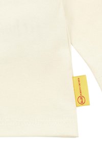 Cremfarbendes Langarmshirt aus weicher Baumwolle. Mit einem gelben Etikett mit rotem Text am Saum. Glatte Textur mit ordentlicher Naht.