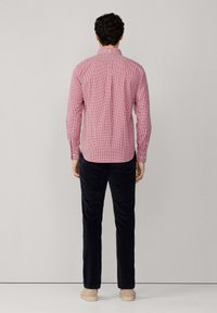 Homme portant une chemise à carreaux rouges à manches longues, un pantalon noir et des chaussures beiges se tenant face à un mur gris clair uni.