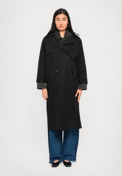 Mujer de pie contra un fondo blanco, vestida con un abrigo negro de doble botonadura largo, jeans azul de pierna ancha y zapatos negros.