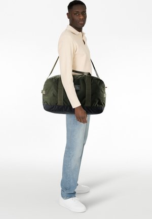 Mann trägt beige Langarmshirt und helle Jeans, trägt große grüne und schwarze Reisetasche über der Schulter, steht vor weißem Hintergrund.