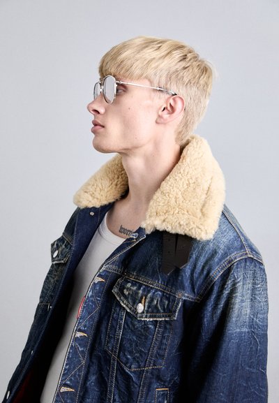 Denim jacket with a fluffy beige collar, front pockets, and black strap detail. Model wearing silver round sunglasses, slight profile view.