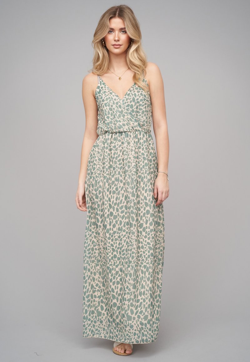 Jeune femme portant une robe sans manches, cintrée à la taille, longue jusqu'aux chevilles, avec des taches abstraites vertes sur fond beige.