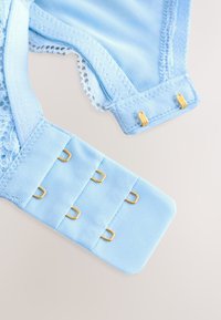 Šviesiai mėlynos spalvos liemenėlės dirželis su nėrinių apdaila; turi lygią juostą su keturiais reguliuojamais kabliukais ir akių užsegimais bei auksinės spalvos aparatūros detalėmis.