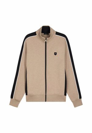 Veste beige zippée avec des rayures noires et blanches sur les manches, un col montant, et une petite écusson logo noir sur la poitrine gauche.