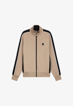 Veste beige zippée avec des rayures noires et blanches sur les manches, un col montant, et une petite écusson logo noir sur la poitrine gauche.