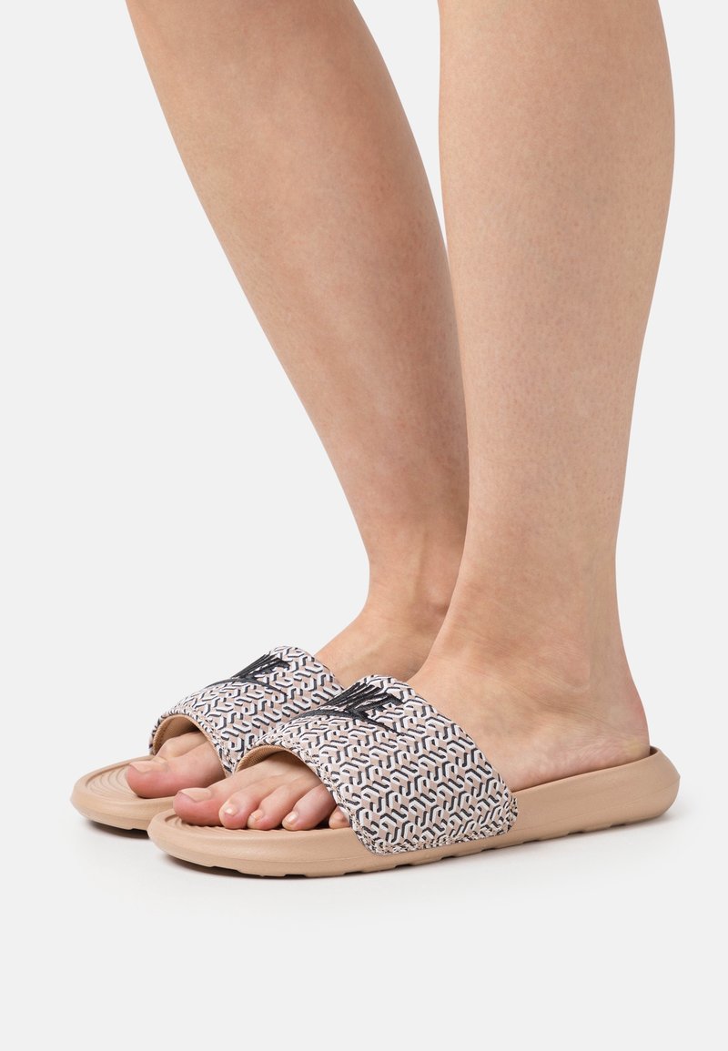 Pieds portant des sandales slides beiges avec larges brides à motifs et logo noir brodé, sur un fond blanc uni.