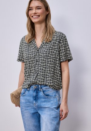 Uśmiechnięta kobieta w czarno-białej bluzce z krótkim rękawem w wzory oraz jasnoniebieskich dżinsach, trzymająca beżową kopertówkę przy boku.