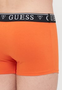 Boxer shorts en coton orange avec une ceinture élastique noire affichant le logo "GUESS" en blanc. Texture lisse, design ajusté.