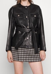 Schwarze Lederjacke mit zwei Brusttaschen, silbernen Knopfakzenten und einem passenden Gürtel. Getragen über einem grauen karierten Minirock.