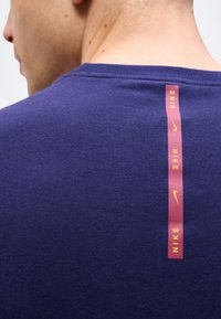 Κοντινό πλάνο ενός ατόμου που φοράει μωβ πουκάμισο με κάθετη κόκκινη λωρίδα που εμφανίζει κίτρινο κείμενο και λογότυπα της Nike στο πάνω μέρος της πλάτης.