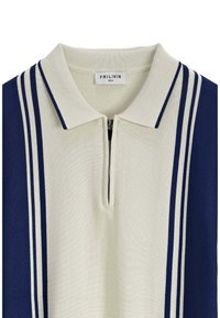 Polo en tricot avec une base crème et des panneaux latéraux navy, trois rayures blanches de chaque côté, fermeture éclair et étiquette de marque visible.