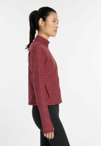 Long-sleeve, deep red athletic top with a mock collar, side slits, and a textured fabric finish, paired with black pants.