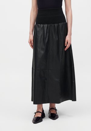 Femme portant une longue jupe en cuir noire avec une large ceinture élastique et des chaussures Mary Jane noires à talons carrés.