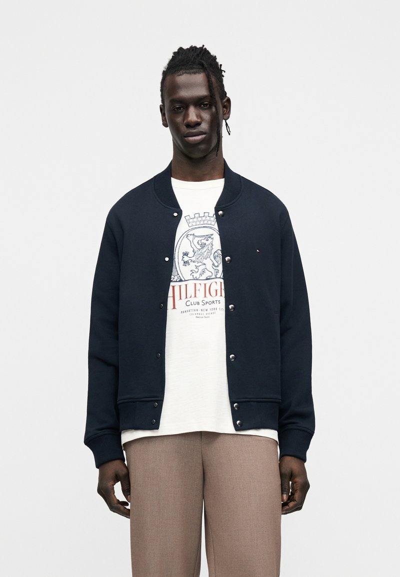 Άνδρας που φοράει ναυτικό μπλε σακάκι με κουμπιά πάνω από λευκό T-shirt με γραφικά και μπεζ παντελόνι, στέκεται μπροστά σε απλό λευκό φόντο.