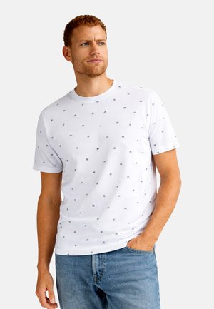 Man met korte krullende haren, gekleed in een wit T-shirt met een kleine donkere opdruk en een blauwe spijkerbroek, kijkt iets opzij met zijn hand in zijn zak.