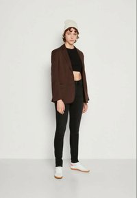 Blazer castanho ajustado sobre um top cropped preto e jeans skinny pretos. Ténis brancos com detalhes laranja. Gorro de malha claro.