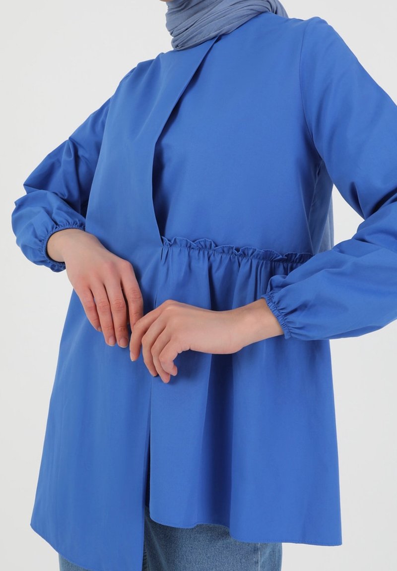 Personne portant un haut bleu à manches longues avec des poignets élastiques et un détail froncé à la taille, associé à un foulard bleu clair et un jean bleu.
