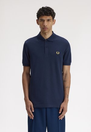 Jeune homme aux cheveux courts et bouclés, portant un polo bleu marine avec un logo laurier jaune et un pantalon assorti, debout droit.